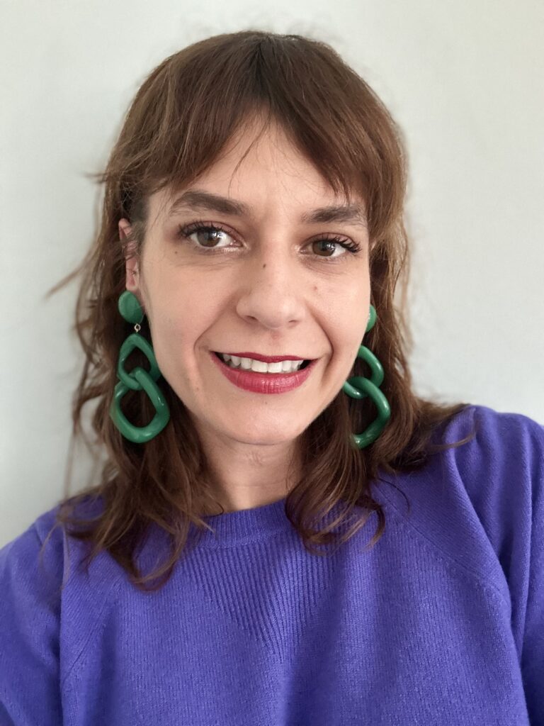 Image of a smiling woman wearing green earrings and a purple jumper