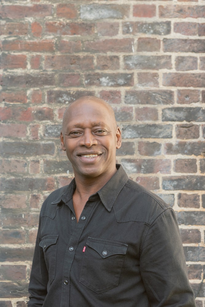 A photographic portrait of a man standing in front of a brick background